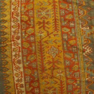 Close-up of a high-pile rug with olive, orange, and gold traditional patterns on distressed wood flooring.