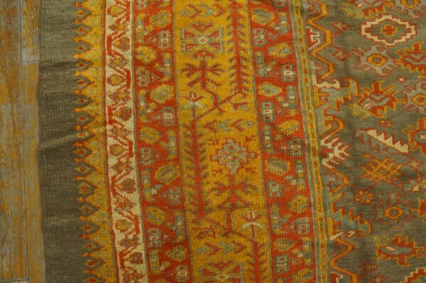 Close-up of a high-pile rug with olive, orange, and gold traditional patterns on distressed wood flooring.