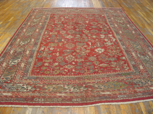 Red traditional rug with intricate motifs and multiple borders, displayed on light brown wood floors.