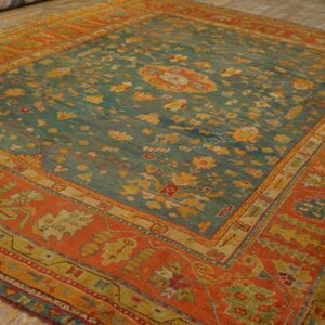 Low-pile rug with a teal floral field and a wide, geometric orange border rests on wood planks.