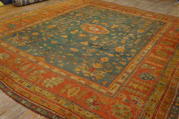Low-pile rug with a teal floral field and a wide, geometric orange border rests on wood planks.