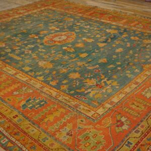 Teal low-pile rug with gold and red motifs, featuring a wide rust-colored patterned border on wood flooring.