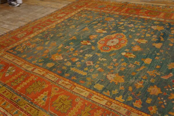 A large rug with a faded teal field and orange-red border is on rustic wood planks.