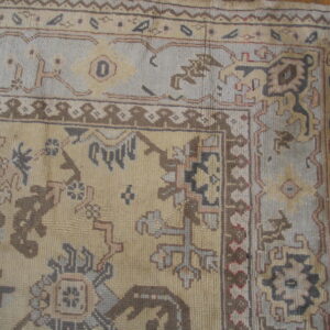 Geometric pattern rug corner in muted beige, blue-gray, and brown with fringed edges on wood.