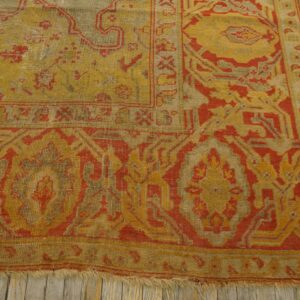 A corner section of a gold and red patterned rug with low pile lying on distressed light wood floors.