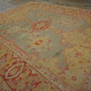 Muted green and gold low-pile rug with red traditional patterns lies on distressed dark wood flooring.