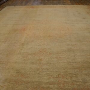 Large beige rug with faded coral patterns lying on a wooden floor near stacks of rolled rugs.