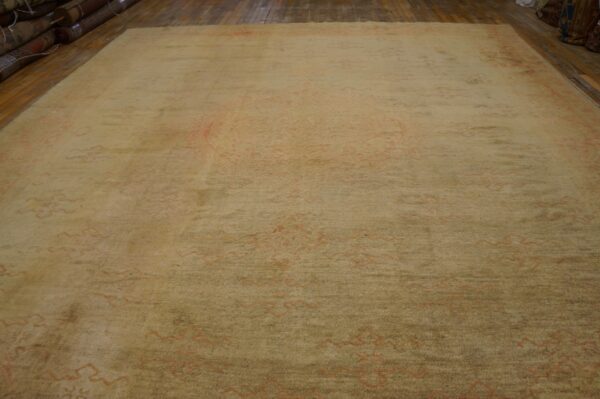 Large beige rug with faded coral patterns lying on a wooden floor near stacks of rolled rugs.