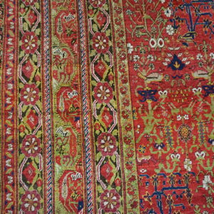 Red rug featuring a detailed floral and vine pattern with striped borders on light brown wood flooring.