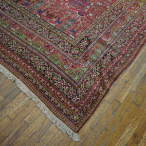 Red ornate rug corner with floral borders and fringe rests on rustic honey-colored wood flooring.