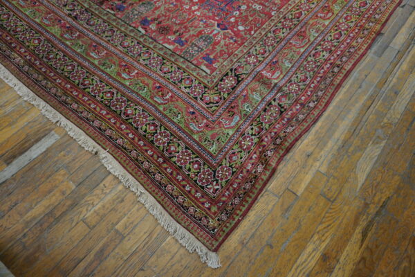 Red ornate rug corner with floral borders and fringe rests on rustic honey-colored wood flooring.