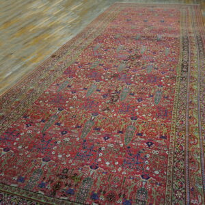 Detailed red rug featuring complex green, blue, and white floral motifs, laid on light plank wood flooring.