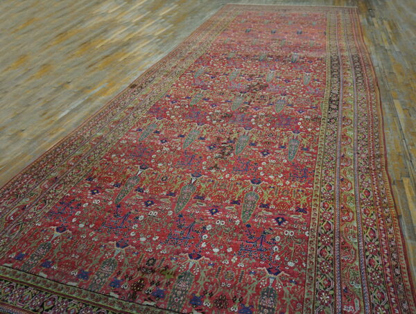 Detailed red rug featuring complex green, blue, and white floral motifs, laid on light plank wood flooring.