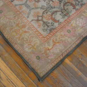 Corner of a low-pile beige rug with rust and grey floral patterns over distressed wood planks.