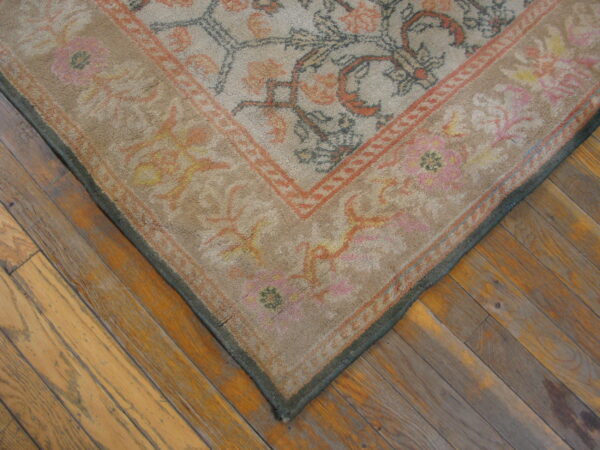 Corner of a low-pile beige rug with rust and grey floral patterns over distressed wood planks.