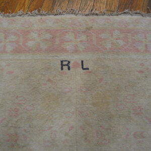 Beige high-pile rug with faded pink borders, black "R L" initials, and fringe rests on hardwood flooring.