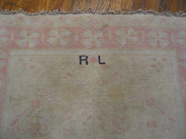 Beige high-pile rug with faded pink borders, black "r l" initials, and fringe rests on hardwood flooring.