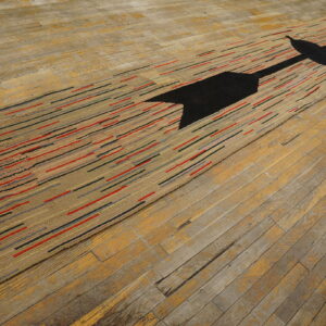 Low-pile rug features a black arrow and tan, red, and gray stripes on worn hardwood floorboards.