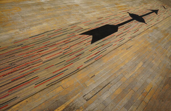 Low-pile rug features a black arrow and tan, red, and gray stripes on worn hardwood floorboards.