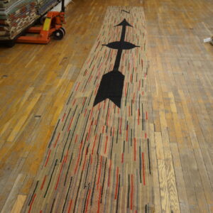 A striped runner rug with a black arrow lies on wood floors near stacked rugs and a pallet jack.