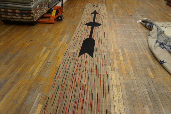 A striped runner rug with a black arrow lies on wood floors near stacked rugs and a pallet jack.
