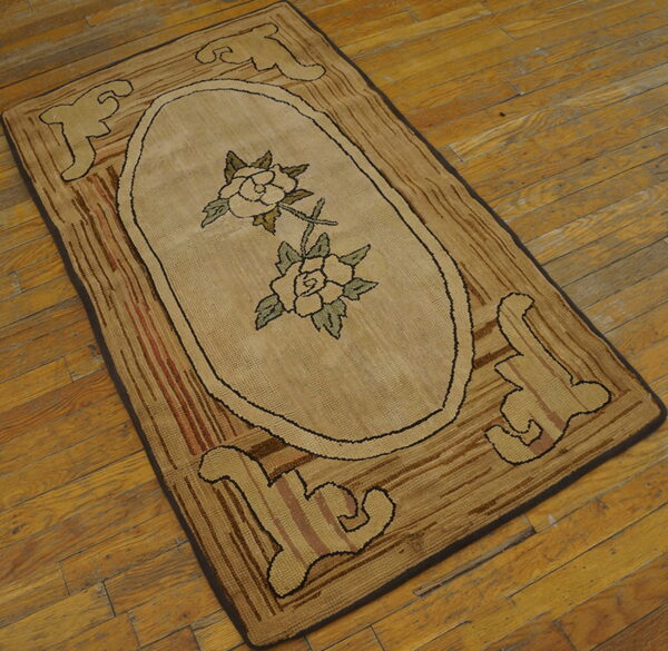 Tan hooked rug with central floral oval and striped brown border, resting on worn hardwood flooring.