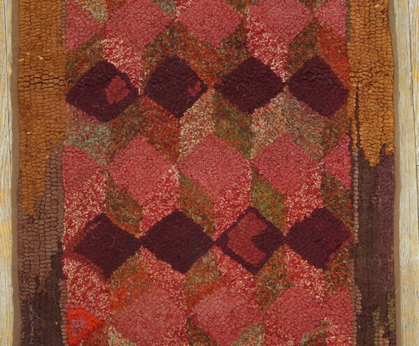 Heavily textured rug featuring a geometric diamond pattern in reds, burgundy, and earth tones against light wood.