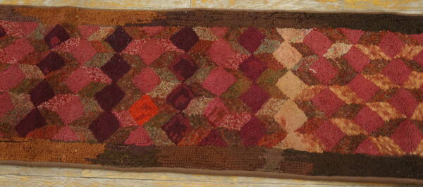 Looped pile rug with a variegated diamond pattern in deep reds, pinks, tans, and dark brown borders on wood flooring.