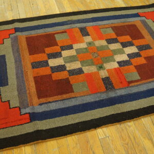 Geometric rug with multi-color blocks and concentric borders of orange, blue, and green on light wood planks.