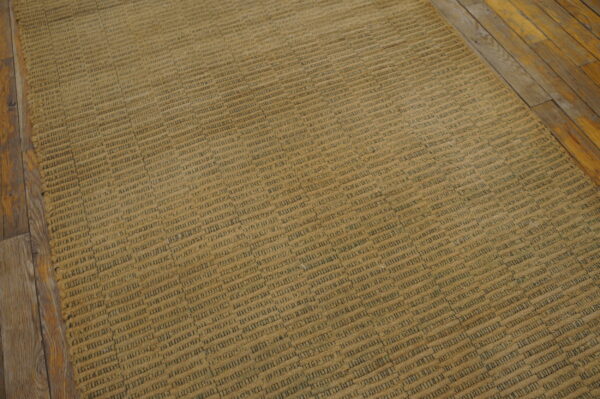 A textured tan rug with dark gray dash patterns lies diagonally on distressed hardwood flooring.