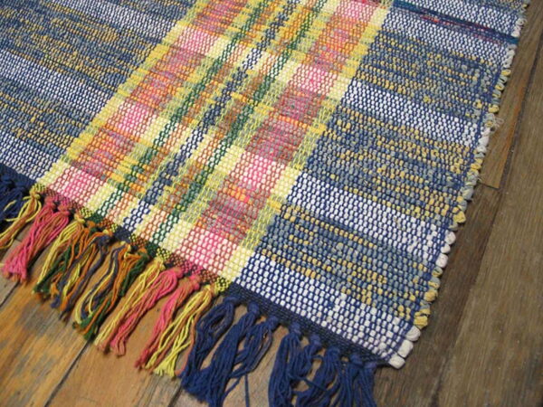 A brightly colored flat-woven plaid rug with blue, pink, and yellow stripes and multicolored tassels rests on a wood floor.