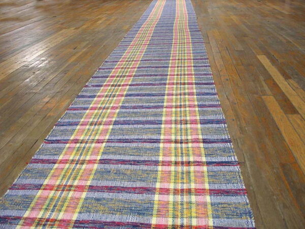 Colorful woven runner rug in blue, pink, and yellow plaid pattern on brown hardwood flooring.