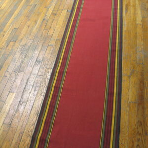A narrow, smooth red runner with striped yellow and green borders lies on a worn wooden plank floor.