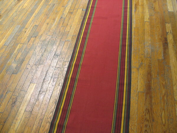 A narrow, smooth red runner with striped yellow and green borders lies on a worn wooden plank floor.