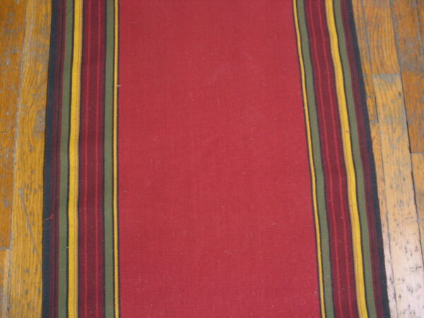 A solid red rug runner with dark multicolored border stripes rests on light hardwood floors.