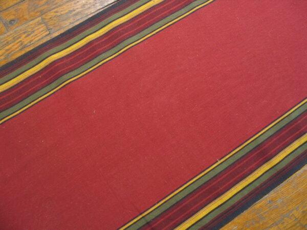 A deep red flatweave rug runner with striped borders in yellow, olive, and burgundy on wood flooring.