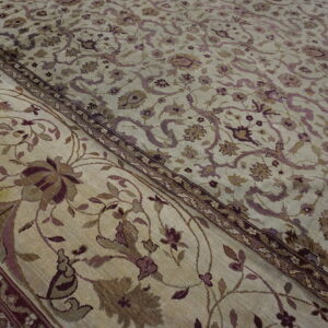 Close-up of a low-pile rug showing a tan field and border with muted purple and brown florals.
