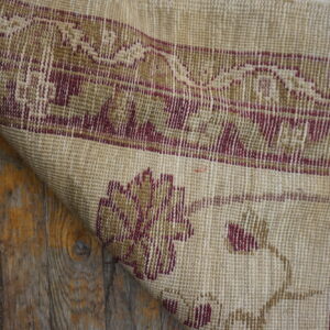 Detail of a tightly woven rug, beige with red and gold patterns, folded over rustic wooden planks.