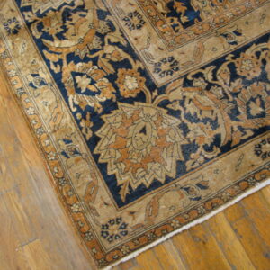 Close-up of a low-pile rug corner with ornate tan and navy blue designs on wood plank flooring.