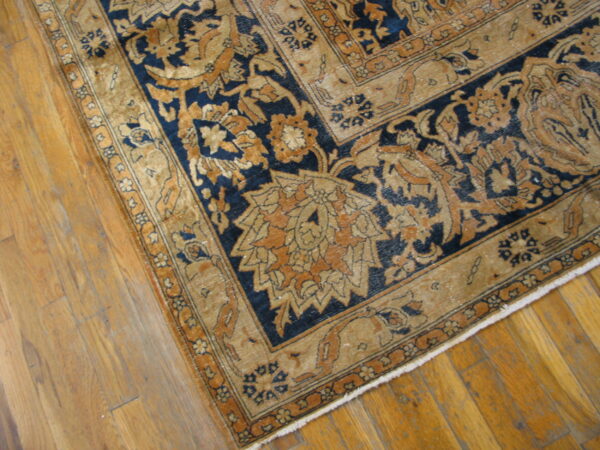 Close-up of a low-pile rug corner with ornate tan and navy blue designs on wood plank flooring.