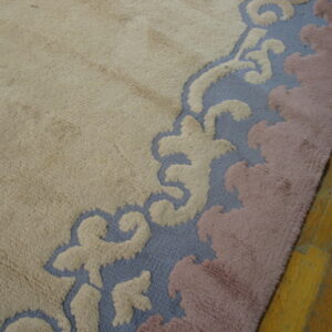 Textured, high-pile rug in beige with a sculpted blue, cream, and mauve scrolled border on wood planks.