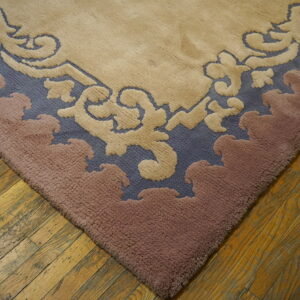 Corner of a carved rug with scrolling motifs in beige, slate blue, and dusty rose on wood planks.