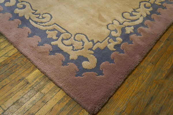 Corner of a carved rug with scrolling motifs in beige, slate blue, and dusty rose on wood planks.