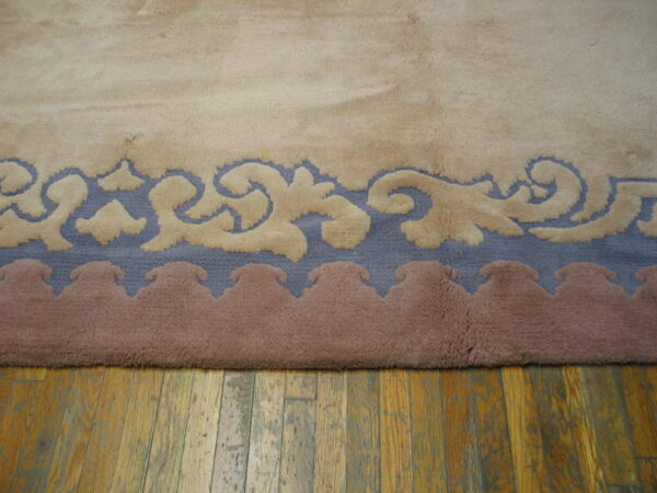 High-pile rug edge featuring beige scrolling patterns on blue and mauve borders over a wood floor.