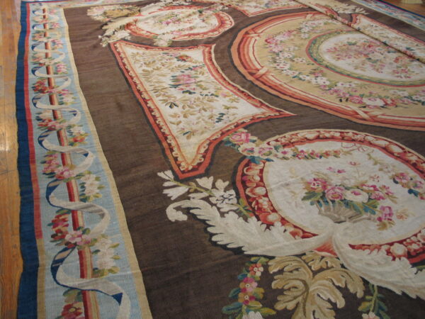 Low-pile rug with complex floral cartouches, cream panels, and a dark brown ribbed field on wood floor.
