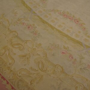 Pale yellow and cream low-pile rug featuring elaborate classical scrollwork, green ribbons, and pink floral details.