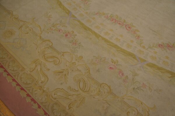 Pale yellow and cream low-pile rug featuring elaborate classical scrollwork, green ribbons, and pink floral details.