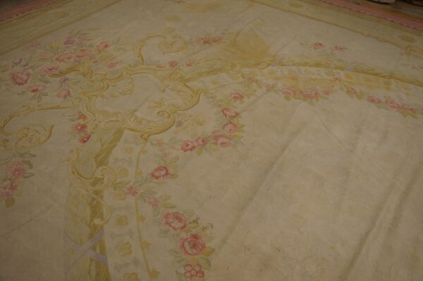 Large, low-pile rug with elaborate scrollwork and floral motifs in pale pink, yellow, and cream.