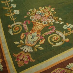 A low-pile green rug displays a floral basket surrounded by brown and colorful scrolling borders.