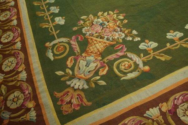 A low-pile green rug displays a floral basket surrounded by brown and colorful scrolling borders.